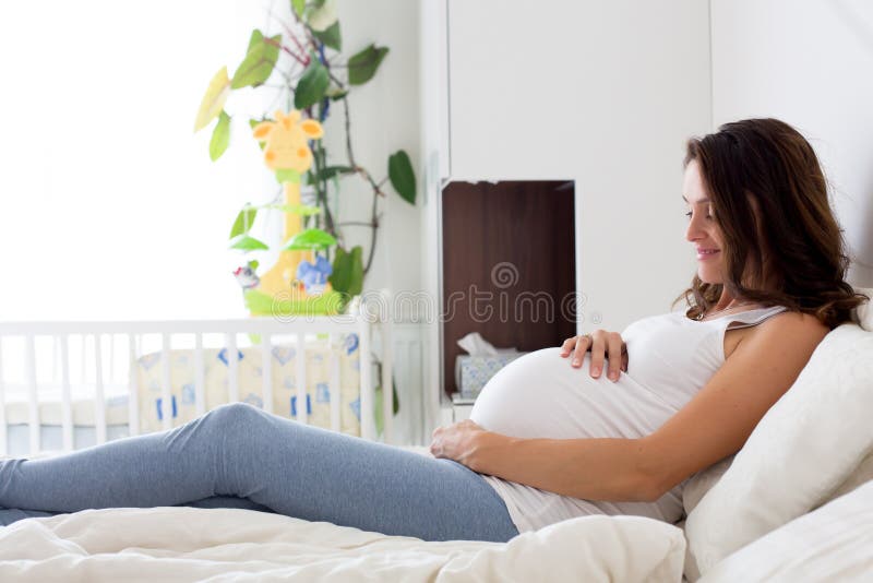 Giovane bella donna incinta felice, sedentesi sul letto in camera da letto fotografia stock libera da diritti Giovane bella donna incinta felice, sedentesi sul letto in camera da letto fotografia stock libera da diritti