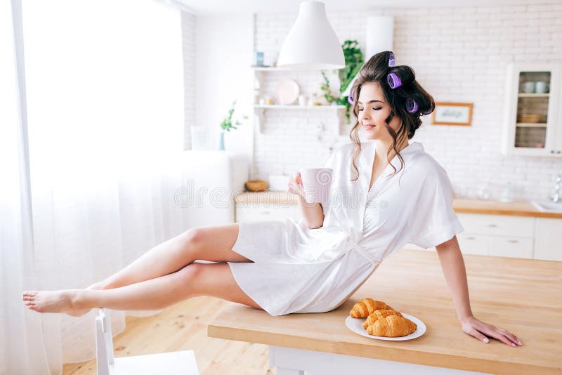 Giovane bella donna spensierata sedersi e riposare sulla tavola in cucina Mattina Castana con i bigodini nel tè della bevanda dei fotografia stock Giovane bella donna spensierata sedersi e riposare sulla tavola in cucina Mattina Castana con i bigodini nel tè della bevanda dei fotografia stock