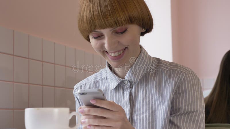 Giovane bella ragazza dai capelli rossi che si siede in un caffè, facendo uso di uno smartphone, mandando un sms, sorridendo, rid