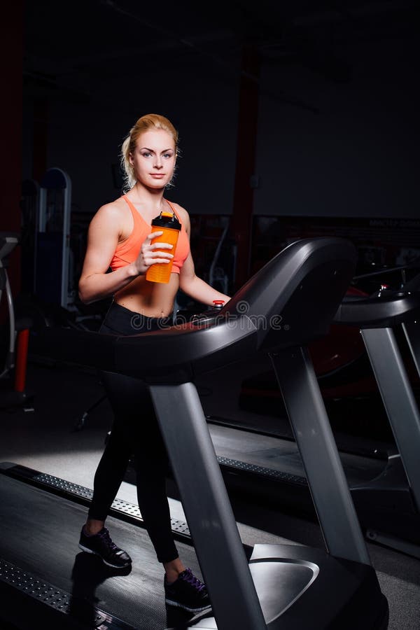 Giovane bella ragazza sveglia nel modello di forma fisica nel funzionamento della palestra sulla pedana mobile con la bottiglia immagini stock Giovane bella ragazza sveglia nel modello di forma fisica nel funzionamento della palestra sulla pedana mobile con la bottiglia immagini stock