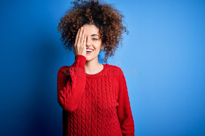 Giovane bellissima donna dai capelli ricci e piercing con un maglione rosso casual che copre un occhio con un sorriso sicuro di ma immagine stock libera da diritti