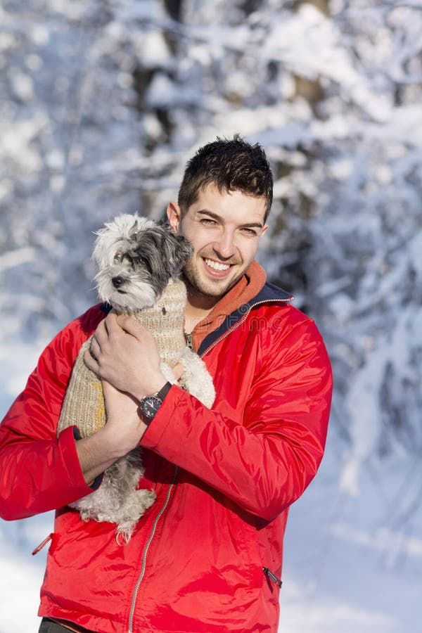 Giovane bello che abbraccia il suo piccolo cane bianco nell'inverno nevicare immagine stock libera da diritti Giovane bello che abbraccia il suo piccolo cane bianco nell'inverno nevicare immagine stock libera da diritti