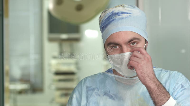 Giovane chirurgo esperto, uomo serio che indossa mascherina da volto, che guarda la telecamera