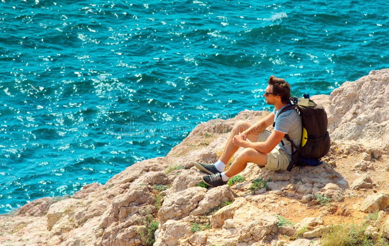 Giovane con lo zaino che si rilassa sulla scogliera rocciosa con il mare blu su fondo fotografia stock Giovane con lo zaino che si rilassa sulla scogliera rocciosa con il mare blu su fondo fotografia stock