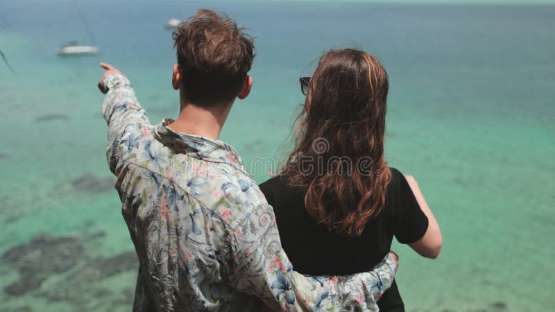 Giovane coppia ammiratrice del paesaggio tropicale con barche a vela all'orizzonte archivi video