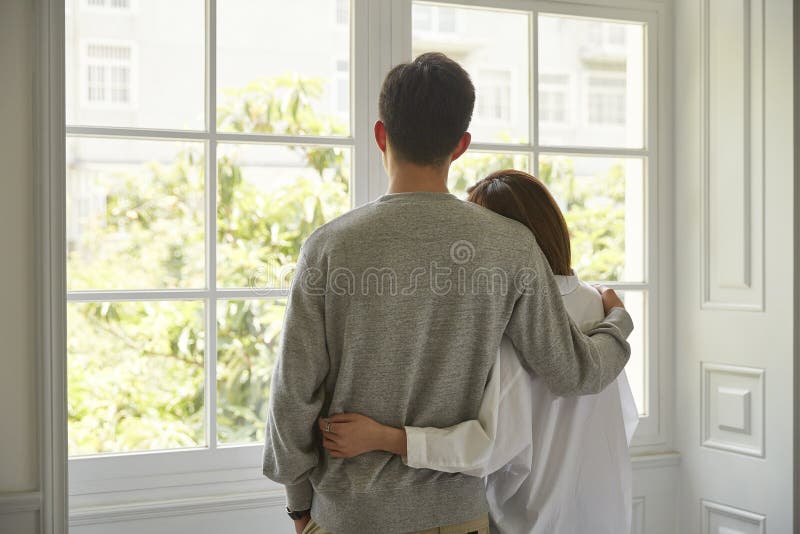 Rear view of a young asian couple looking out of window during staying at home order. Rear view of a young asian couple looking out of window during staying at home order