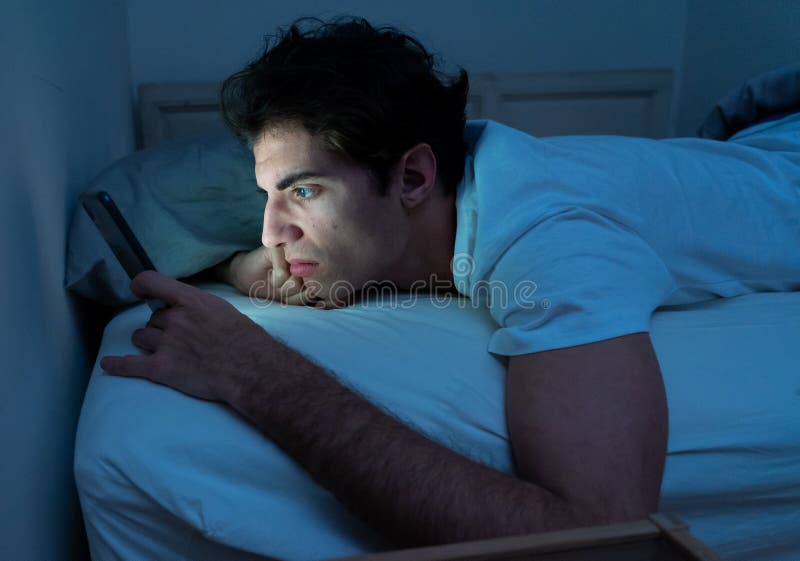 Addicted to social media young man chatting and surfing on the Internet on smart phone at night in bed. Sleepless in dark bedroom with mobile screen light. In insomnia and online network addiction. Addicted to social media young man chatting and surfing on the Internet on smart phone at night in bed. Sleepless in dark bedroom with mobile screen light. In insomnia and online network addiction