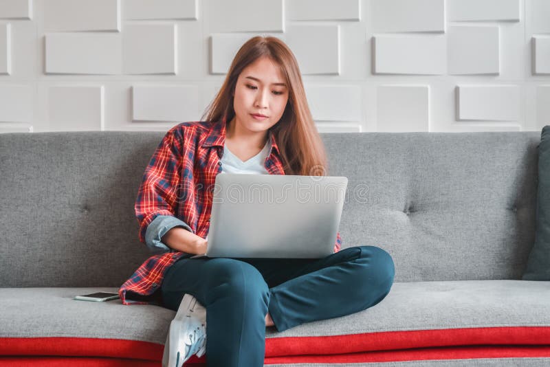 Giovane donna in abbigliamento casual facendo uso del computer portatile che si siede funzionamento rilassato sullo strato domest fotografia stock Giovane donna in abbigliamento casual facendo uso del computer portatile che si siede funzionamento rilassato sullo strato domest fotografia stock