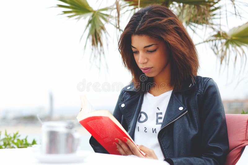 Young lovely woman sitting at coffee shop terrace pensive reading interesting book, afro american female enjoying a good book or novel during her recreation time outdoors at weekend. Young lovely woman sitting at coffee shop terrace pensive reading interesting book, afro american female enjoying a good book or novel during her recreation time outdoors at weekend