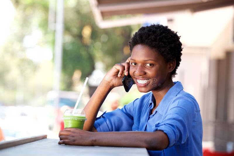 Giovane donna africana sorridente che si siede al caffè ed a fare telefonata immagini stock Giovane donna africana sorridente che si siede al caffè ed a fare telefonata immagini stock