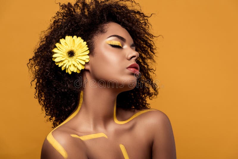 Giovane donna afroamericana attraente con trucco artistico e gerbera in capelli immagine stock Giovane donna afroamericana attraente con trucco artistico e gerbera in capelli immagine stock