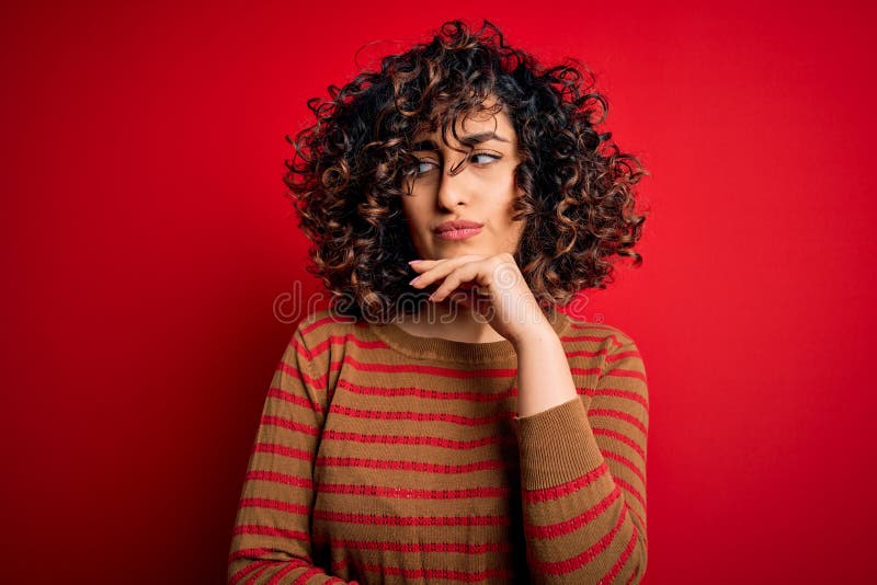 Giovane donna araba ricciola con un maglione casual a strisce in piedi sullo sfondo rosso con mano sul mento pensando immagini stock libere da diritti