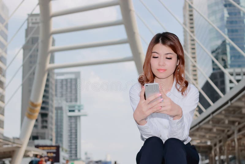 Giovane donna asiatica attraente in camicia bianca che osserva Smart Phone mobile in sue mani il fondo di costruzione urbano Vita immagini stock Giovane donna asiatica attraente in camicia bianca che osserva Smart Phone mobile in sue mani il fondo di costruzione urbano Vita immagini stock