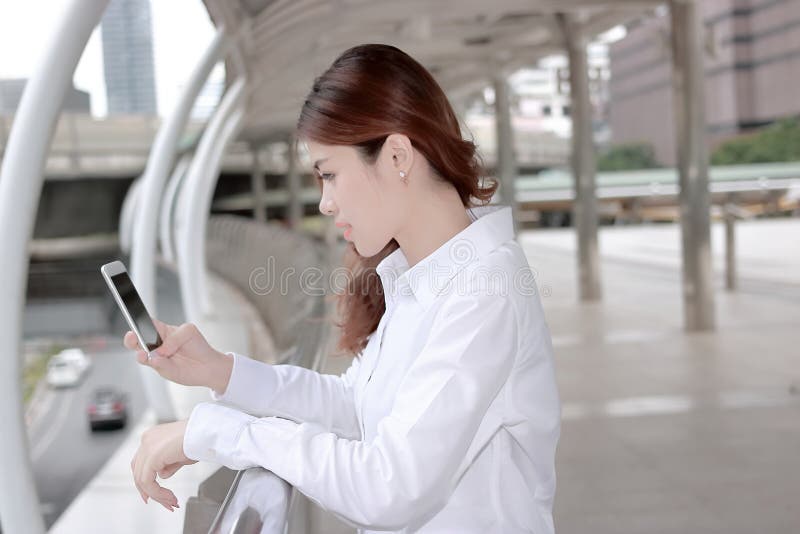 Giovane donna asiatica attraente che considera Smart Phone mobile in sue mani a sviluppare fondo urbano fotografia stock libera da diritti Giovane donna asiatica attraente che considera Smart Phone mobile in sue mani a sviluppare fondo urbano fotografia stock libera da diritti