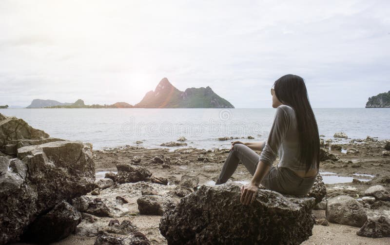 Giovane donna asiatica che si siede su una roccia vicino al mare, guardato al mare, al freddo da estate, al tempo di resto, alla immagini stock Giovane donna asiatica che si siede su una roccia vicino al mare, guardato al mare, al freddo da estate, al tempo di resto, alla immagini stock