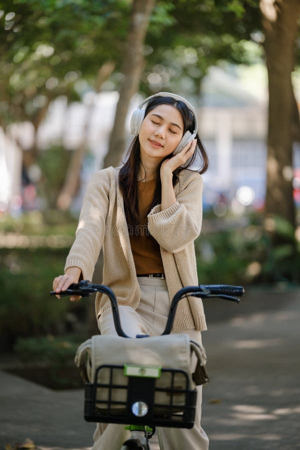 Giovane donna asiatica usa cuffie mentre va in bicicletta nel parco cittadino fotografia stock libera da diritti