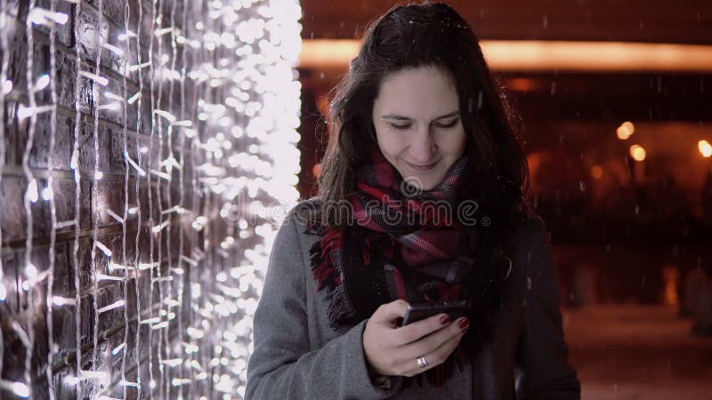 Giovane donna attraente che parla sul telefono nella neve di caduta alla notte di Natale che sta la parete vicina delle luci, immagini stock libere da diritti Giovane donna attraente che parla sul telefono nella neve di caduta alla notte di Natale che sta la parete vicina delle luci, immagini stock libere da diritti