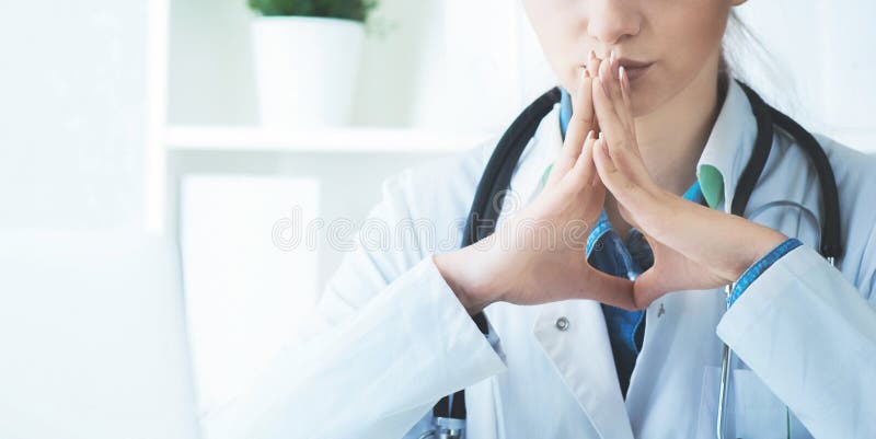Giovane donna bionda seria di medico che si siede nell'ufficio medico con le mani davanti al fronte che pensa alla domanda, pensi immagine stock libera da diritti Giovane donna bionda seria di medico che si siede nell'ufficio medico con le mani davanti al fronte che pensa alla domanda, pensi immagine stock libera da diritti