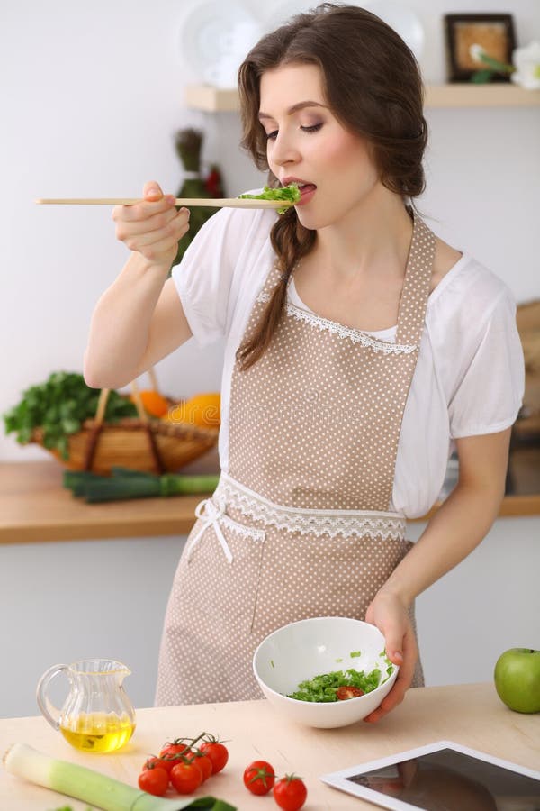 Giovane donna castana che cucina nella cucina Casalinga che tiene cucchiaio di legno in sua mano Alimento e concetto di salute immagini stock libere da diritti Giovane donna castana che cucina nella cucina Casalinga che tiene cucchiaio di legno in sua mano Alimento e concetto di salute immagini stock libere da diritti