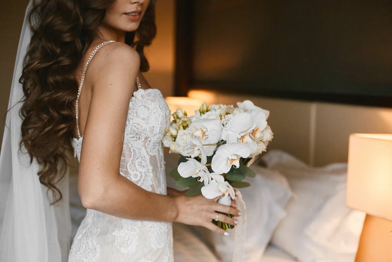 Giovane donna castana con l'acconciatura di nozze in un vestito da sposa dal pizzo con un mazzo dei fiori freschi in sue mani den fotografia stock libera da diritti Giovane donna castana con l'acconciatura di nozze in un vestito da sposa dal pizzo con un mazzo dei fiori freschi in sue mani den fotografia stock libera da diritti
