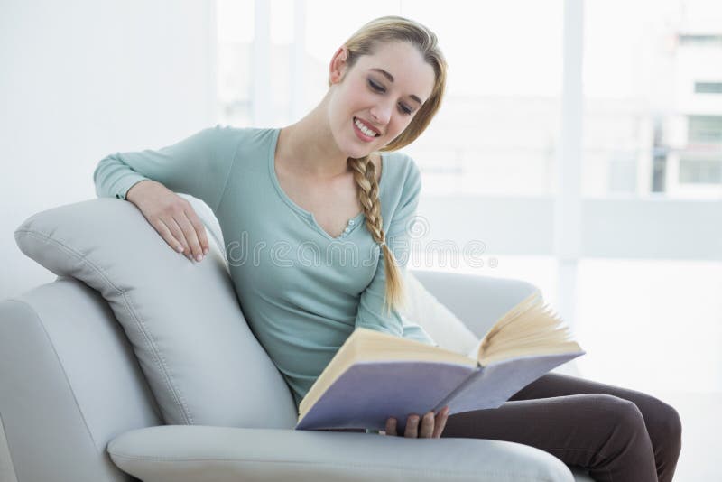 Giovane donna casuale che legge un libro mentre sedendosi sorridere sullo strato immagini stock libere da diritti Giovane donna casuale che legge un libro mentre sedendosi sorridere sullo strato immagini stock libere da diritti