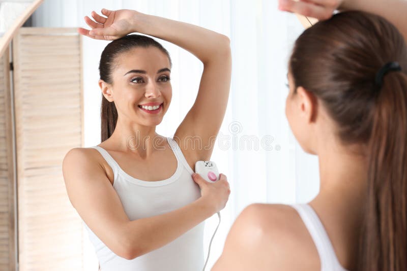 Giovane donna che fa procedura di epilation dell'ascella vicino allo specchio a fotografia stock Giovane donna che fa procedura di epilation dell'ascella vicino allo specchio a fotografia stock