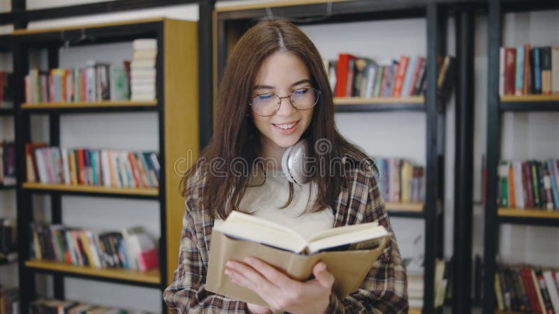 Giovane donna che legge un libro mentre si gode un momento tranquillo in una biblioteca piena di scaffali di romanzi e letteratura video d archivio