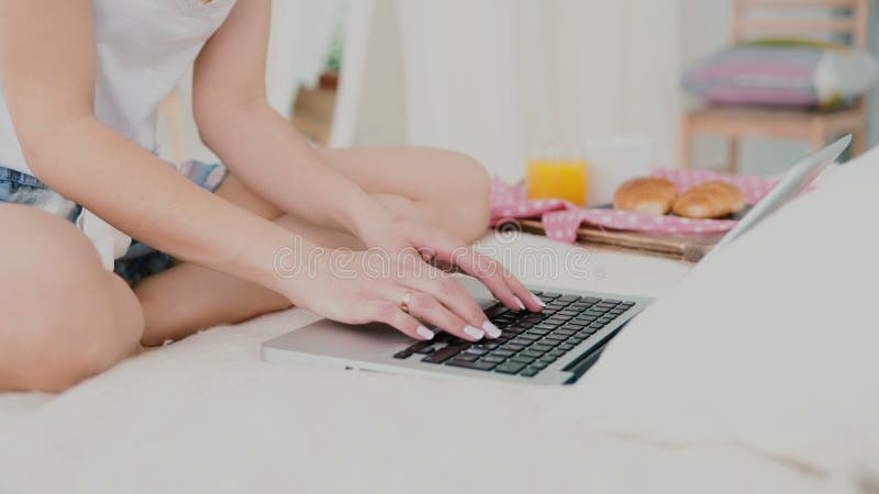 Giovane donna che per mezzo del computer portatile durante la prima colazione che si siede sul letto bianco Vista del primo piano immagini stock libere da diritti Giovane donna che per mezzo del computer portatile durante la prima colazione che si siede sul letto bianco Vista del primo piano immagini stock libere da diritti