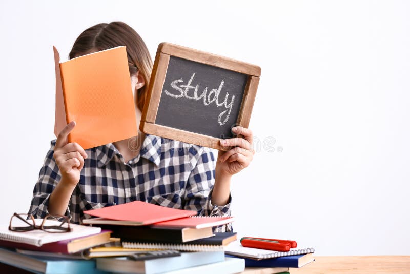 Giovane donna che si siede alla tavola con molti libri e che tiene lavagna con lo studio della parola contro il fondo bianco fotografie stock Giovane donna che si siede alla tavola con molti libri e che tiene lavagna con lo studio della parola contro il fondo bianco fotografie stock