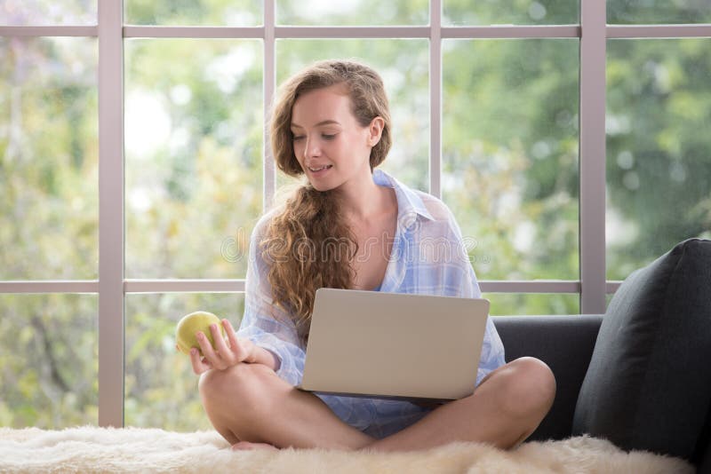 Giovane donna che si siede su uno strato facendo uso del computer portatile e che tiene una mela immagini stock libere da diritti Giovane donna che si siede su uno strato facendo uso del computer portatile e che tiene una mela immagini stock libere da diritti