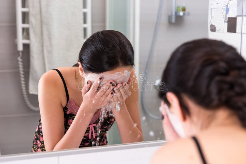 Young woman with cosmetic mask spraying water on her face standing in front of mirror in the modern tiled bathroom at home. Young woman with cosmetic mask spraying water on her face standing in front of mirror in the modern tiled bathroom at home.