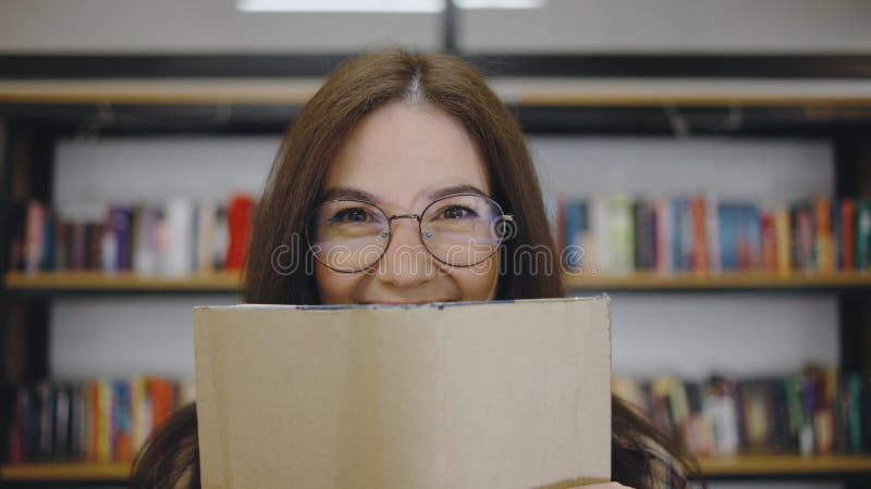 Giovane donna con gli occhiali che nasconde scherzosamente il viso dietro un libro in una comoda biblioteca circondata da scaffali stock footage