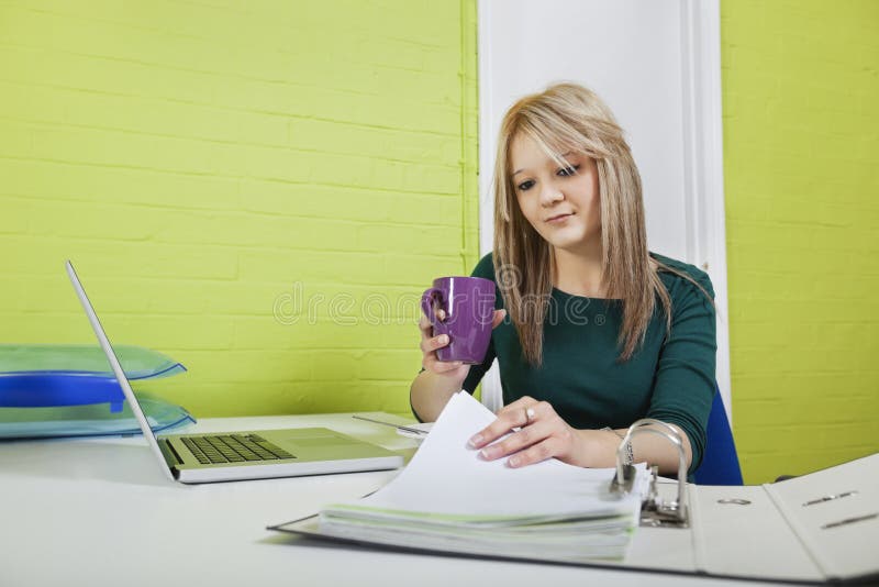 Giovane donna di affari con la tazza da caffè che esamina i documenti allo scrittorio immagini stock