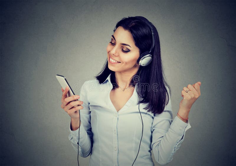 Giovane donna di dancing che ascolta la musica in cuffie e che tiene telefono cellulare su fondo grigio scuro fotografie stock libere da diritti Giovane donna di dancing che ascolta la musica in cuffie e che tiene telefono cellulare su fondo grigio scuro fotografie stock libere da diritti