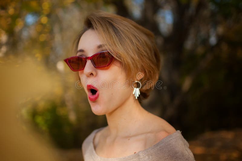 Giovane donna emotivamente attraente con capelli biondi corti, abito da sera e occhiali da sole rossi e fissabili che grimeggiano fotografie stock Giovane donna emotivamente attraente con capelli biondi corti, abito da sera e occhiali da sole rossi e fissabili che grimeggiano fotografie stock