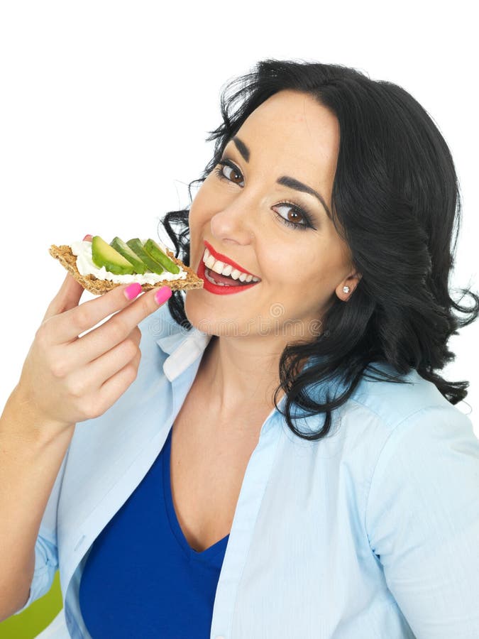 Healthy attractive young woman eating a wholegrain cracker with cottage cheese and fresh avocado slices looking towards the camera smiling healthily, a healthy snack, healthy lunch, brunch, diet or dieting healthy lifestyle food, low calorie, low fat, with wavy dark hair, wearing a blue top with a light blue open shirt or blouse on top, isolated against a white background, clipping path or cut out. Healthy attractive young woman eating a wholegrain cracker with cottage cheese and fresh avocado slices looking towards the camera smiling healthily, a healthy snack, healthy lunch, brunch, diet or dieting healthy lifestyle food, low calorie, low fat, with wavy dark hair, wearing a blue top with a light blue open shirt or blouse on top, isolated against a white background, clipping path or cut out