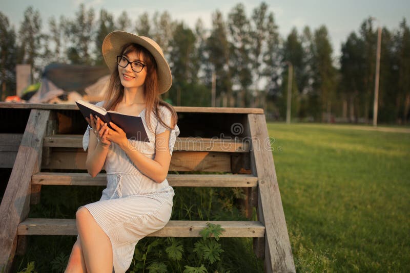 Giovane donna felice che si siede nel parco Sorridendo e tenendo un libro in sue mani sedendosi su un banco di legno Luce di tram immagine stock libera da diritti Giovane donna felice che si siede nel parco Sorridendo e tenendo un libro in sue mani sedendosi su un banco di legno Luce di tram immagine stock libera da diritti