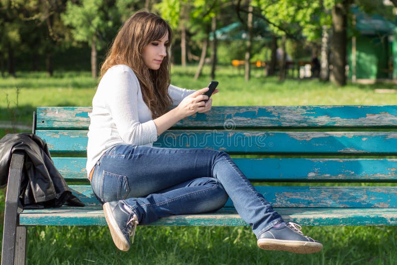 Giovane donna graziosa che per mezzo dello smartphone che si siede sul banco immagini stock Giovane donna graziosa che per mezzo dello smartphone che si siede sul banco immagini stock