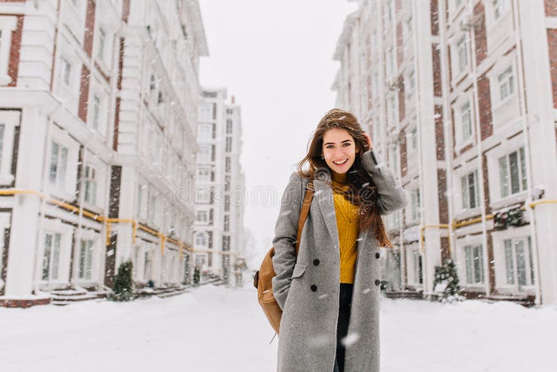 Giovane donna incantante in cappotto con capelli castana lunghi che gode delle precipitazioni nevose in grande città Emozioni all fotografia stock libera da diritti Giovane donna incantante in cappotto con capelli castana lunghi che gode delle precipitazioni nevose in grande città Emozioni all fotografia stock libera da diritti
