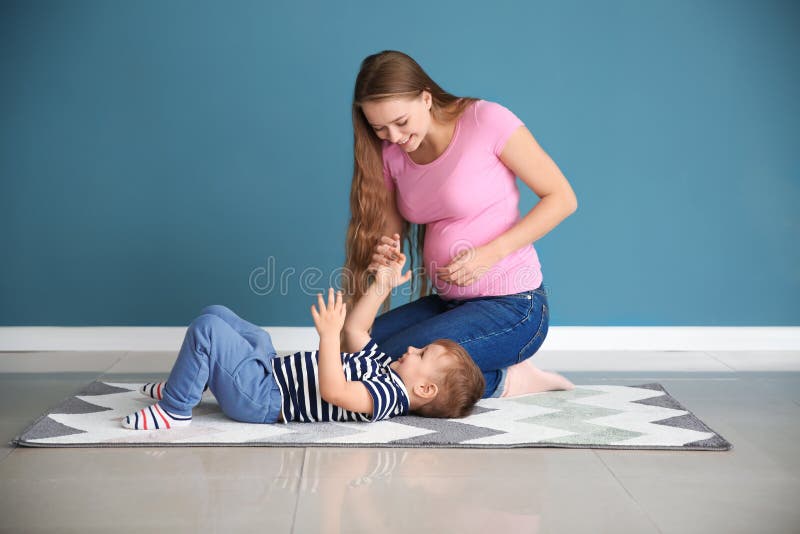 Giovane donna incinta che gioca con il suo sveglio poco figlio su tappeto vicino alla parete di colore fotografie stock libere da diritti Giovane donna incinta che gioca con il suo sveglio poco figlio su tappeto vicino alla parete di colore fotografie stock libere da diritti