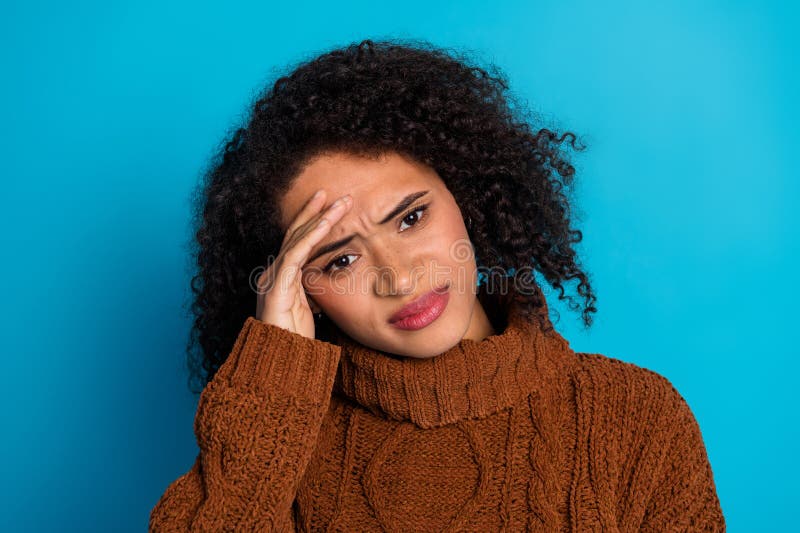 Giovane donna maglione a maglia marrone con la mano sulla testa che esprime confusione sullo sfondo blu. Una giovane donna con un maglione a maglia marrone stringe la mano al tempio esprimendo confusione o sconsideratezza. La scena si svolge su uno sfondo blu chiaro.