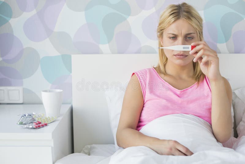 Giovane donna malata che prende temperatura con il termometro a letto immagini stock Giovane donna malata che prende temperatura con il termometro a letto immagini stock