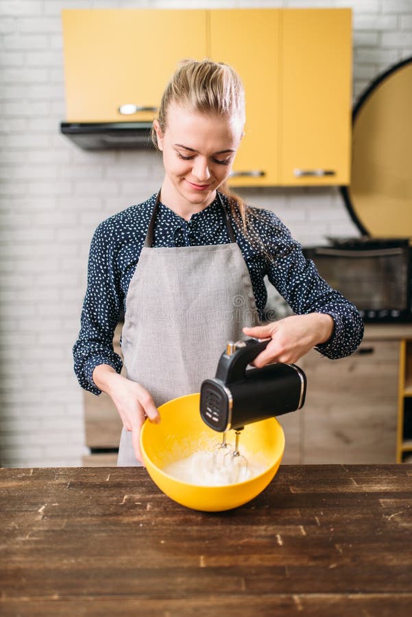 Young woman in apron whip dough in bowl with a mixer. Sweet cake cooking preparation. Kitchen on background. Young woman in apron whip dough in bowl with a mixer. Sweet cake cooking preparation. Kitchen on background