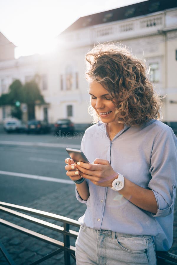 Giovane donna riccia felice che manda un sms e che sorride sullo Smart Phone fotografie stock libere da diritti Giovane donna riccia felice che manda un sms e che sorride sullo Smart Phone fotografie stock libere da diritti