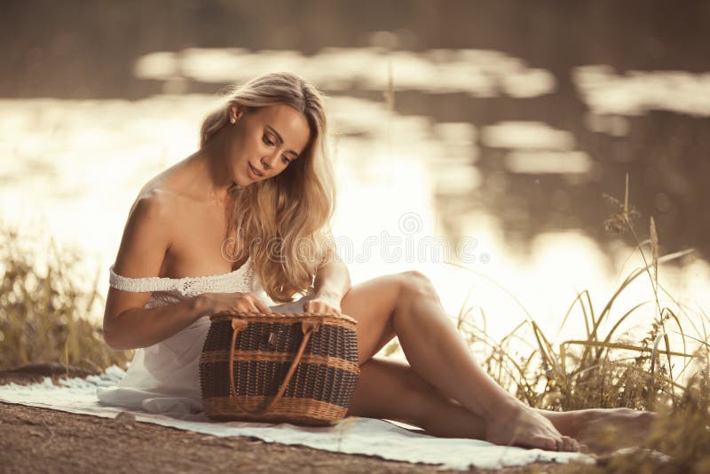 Giovane donna sensuale sul picnic che si siede dal lago al tramonto e che guarda nel canestro di picnic fotografia stock Giovane donna sensuale sul picnic che si siede dal lago al tramonto e che guarda nel canestro di picnic fotografia stock