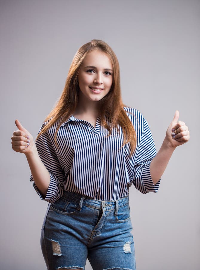 Giovane donna sorridente allegra che mostra i pollici su su fondo grigio e che esamina la macchina fotografica immagini stock Giovane donna sorridente allegra che mostra i pollici su su fondo grigio e che esamina la macchina fotografica immagini stock