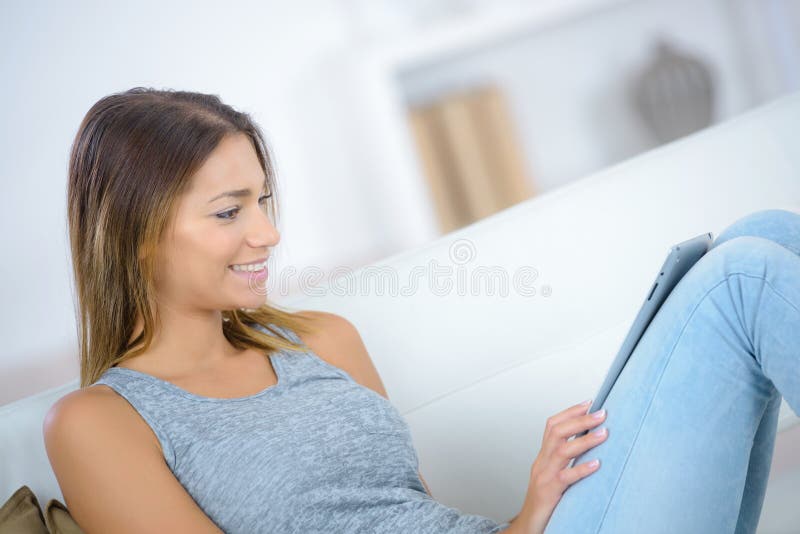 Giovane donna sorridente che si siede sul sofà con la compressa immagine stock libera da diritti Giovane donna sorridente che si siede sul sofà con la compressa immagine stock libera da diritti