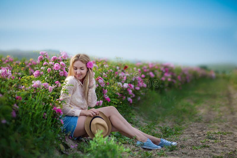 Giovane donna tenera graziosa che cammina nel campo delle rose di tè I jeans d'uso di signora bionda ed il retro cappello godono fotografie stock libere da diritti Giovane donna tenera graziosa che cammina nel campo delle rose di tè I jeans d'uso di signora bionda ed il retro cappello godono fotografie stock libere da diritti