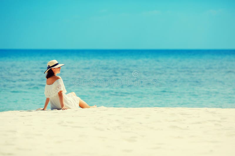 Giovane donna in vestito bianco e cappello che si trovano sulla sabbia della spiaggia tropicale che ha grande ora legale fotografie stock Giovane donna in vestito bianco e cappello che si trovano sulla sabbia della spiaggia tropicale che ha grande ora legale fotografie stock