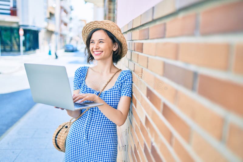 Giovane e bella ragazza che sorride felice usando un portatile appoggiato sul muro per strada fotografie stock